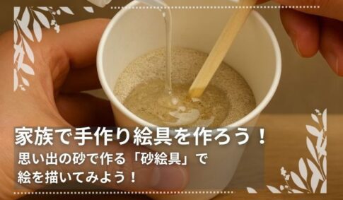 紙コップに入った細かい砂にアラビアゴムメディウムを加え、割りばしで混ぜて絵具を作っている様子。“家族で手作り絵具を作ろう！思い出の砂で作る『砂絵具』で絵を描いてみよう！”というメッセージ入り。